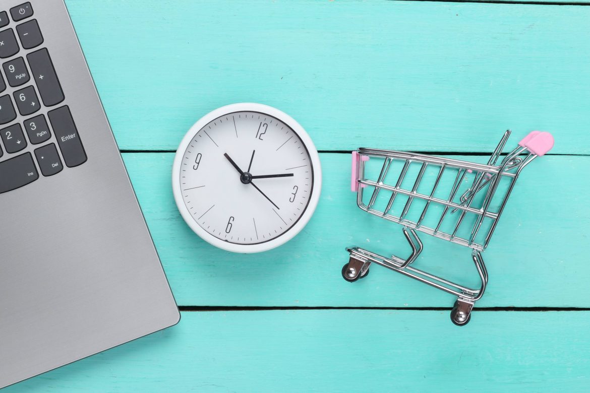 Online shopping time. Computer keyboard with clock and supermarket trolley on blue wooden background. Top view dorzuć do paczki