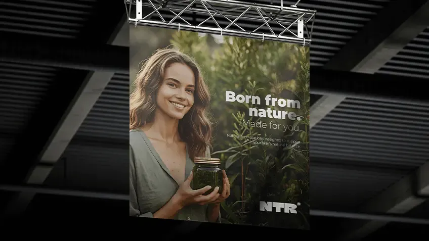 Podwieszany baner tekstylny NTR z hasłem Born from nature. Made for you. oraz wizerunkiem uśmiechniętej kobiety trzymającej słoik z naturalnym produktem